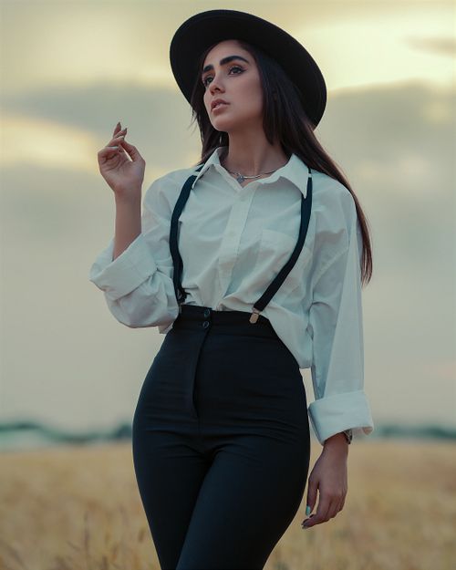 Free Fashion portrait of a sophisticated woman in a field at dusk, wearing a white shirt, hat, and suspenders. Stock Photo