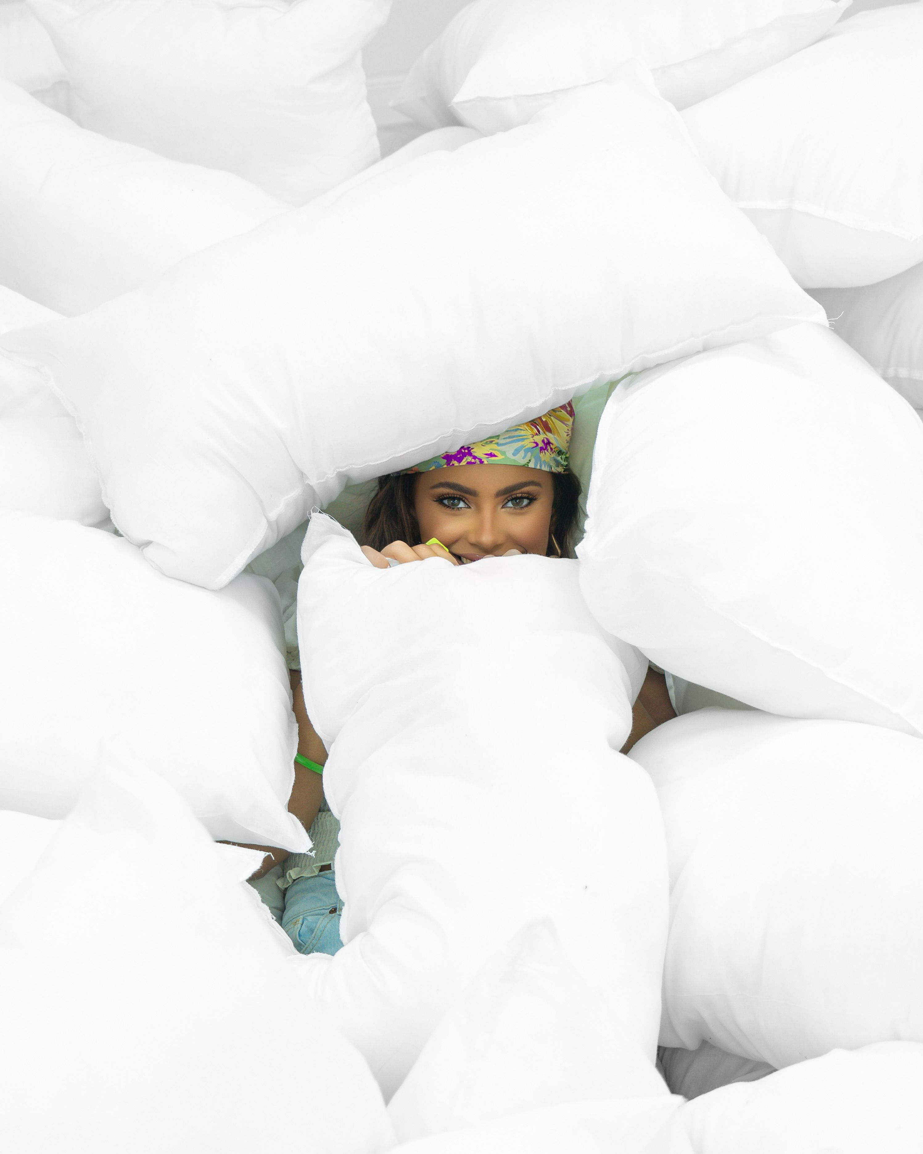 Woman playfully peeking through a stack of fluffy white pillows, creating a fun and cozy atmosphere.
