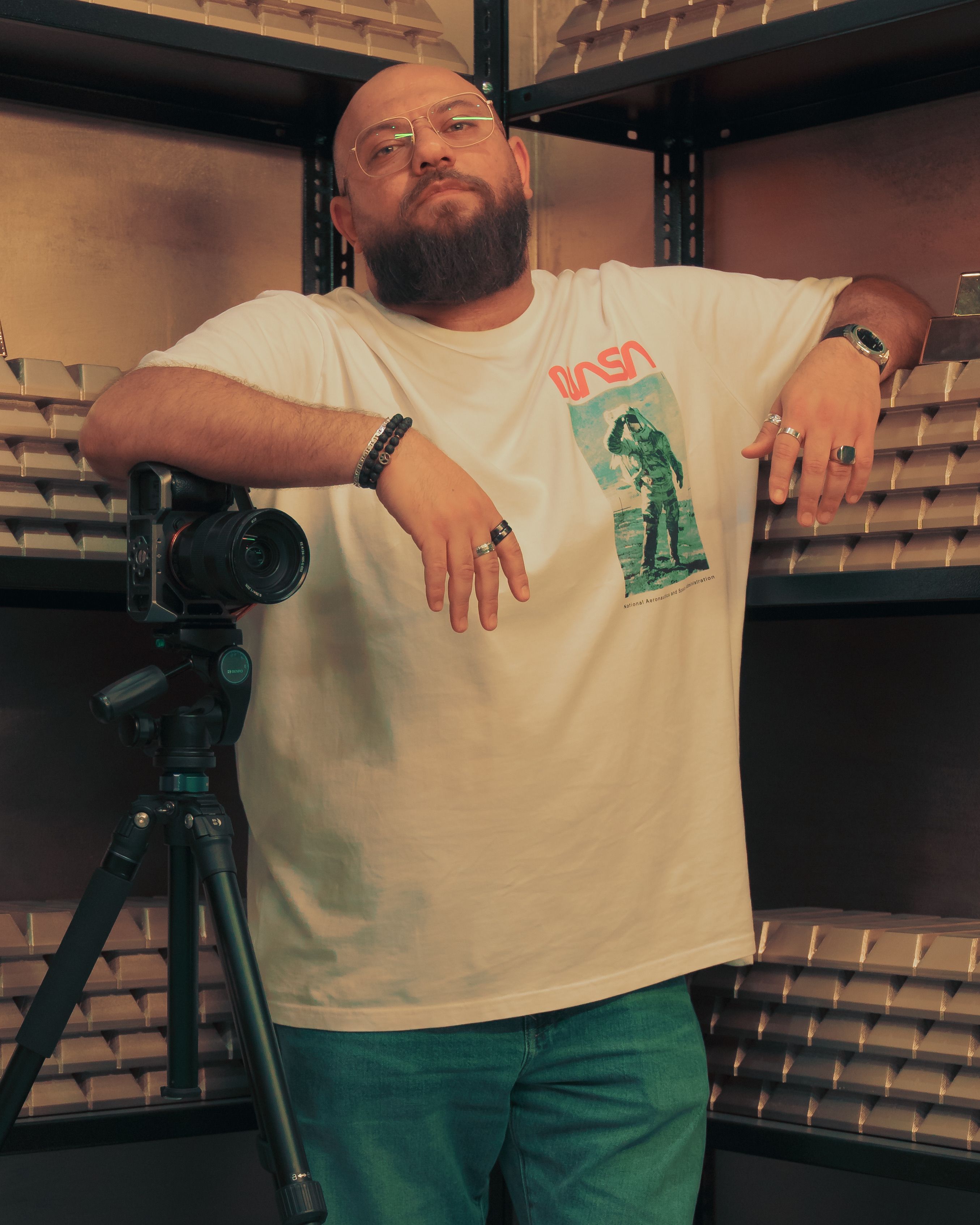 Bald photographer in a white t-shirt with a camera on a tripod poses indoors.
