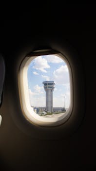 Airplane window view of an airport control tower against a blue sky with clouds.