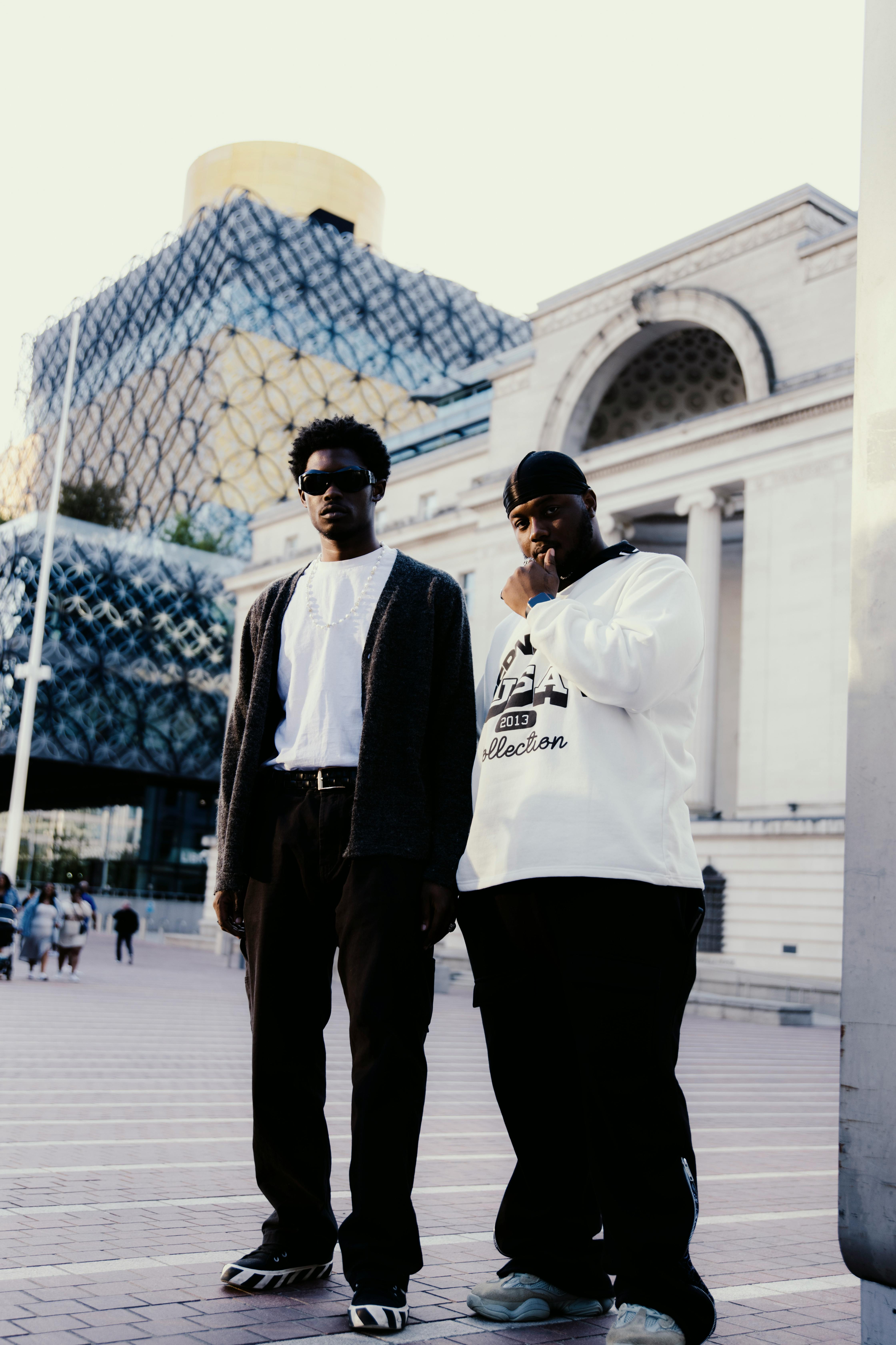 Two men posing in front of Birmingham's Library with modern architecture backdrop.