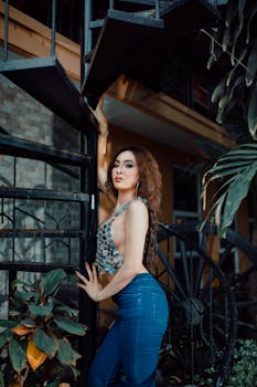 Model in silver top and jeans posing on spiral staircase outdoors with foliage.