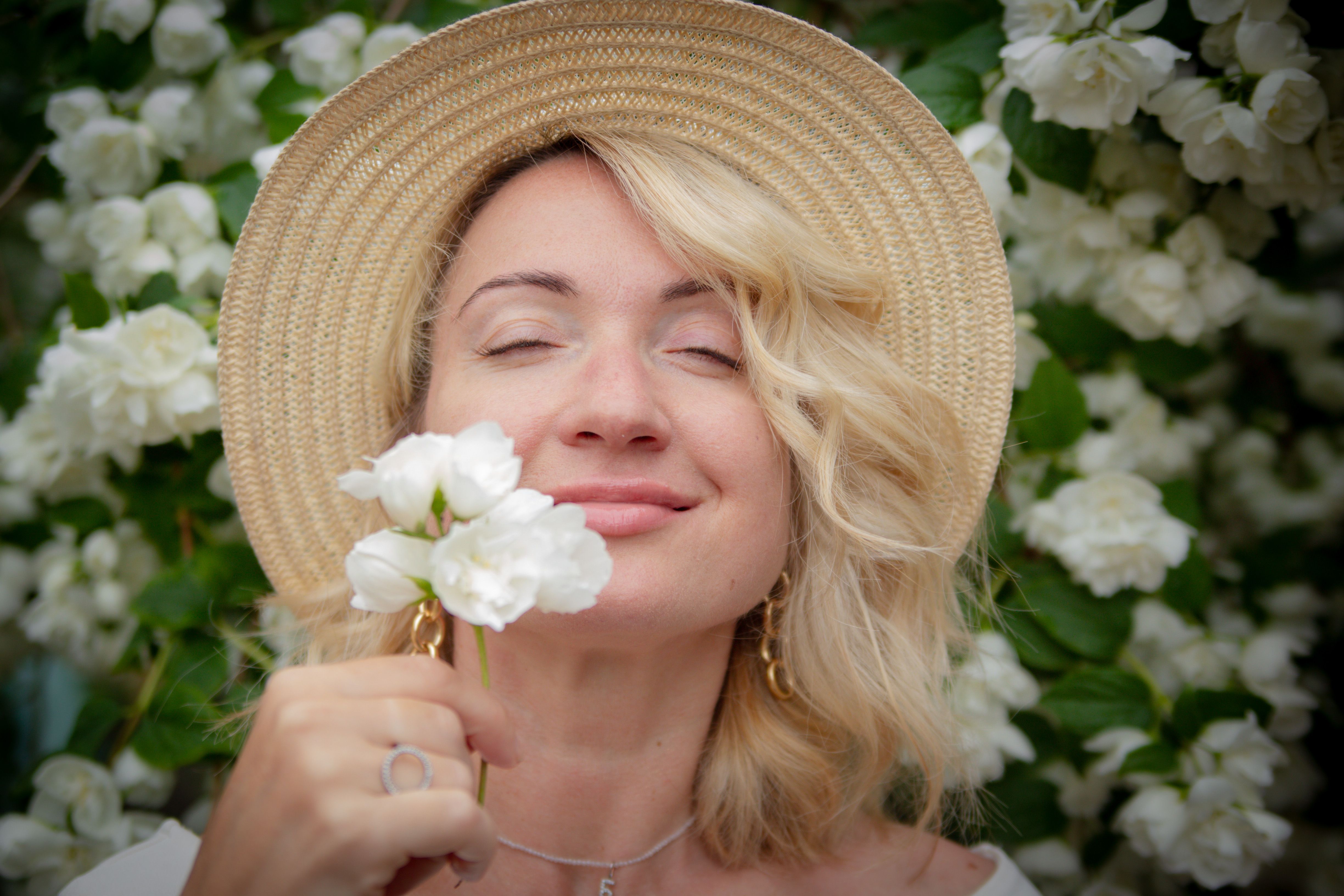 Blonde woman enjoying white flowers outdoors, smiling happily in a straw hat.