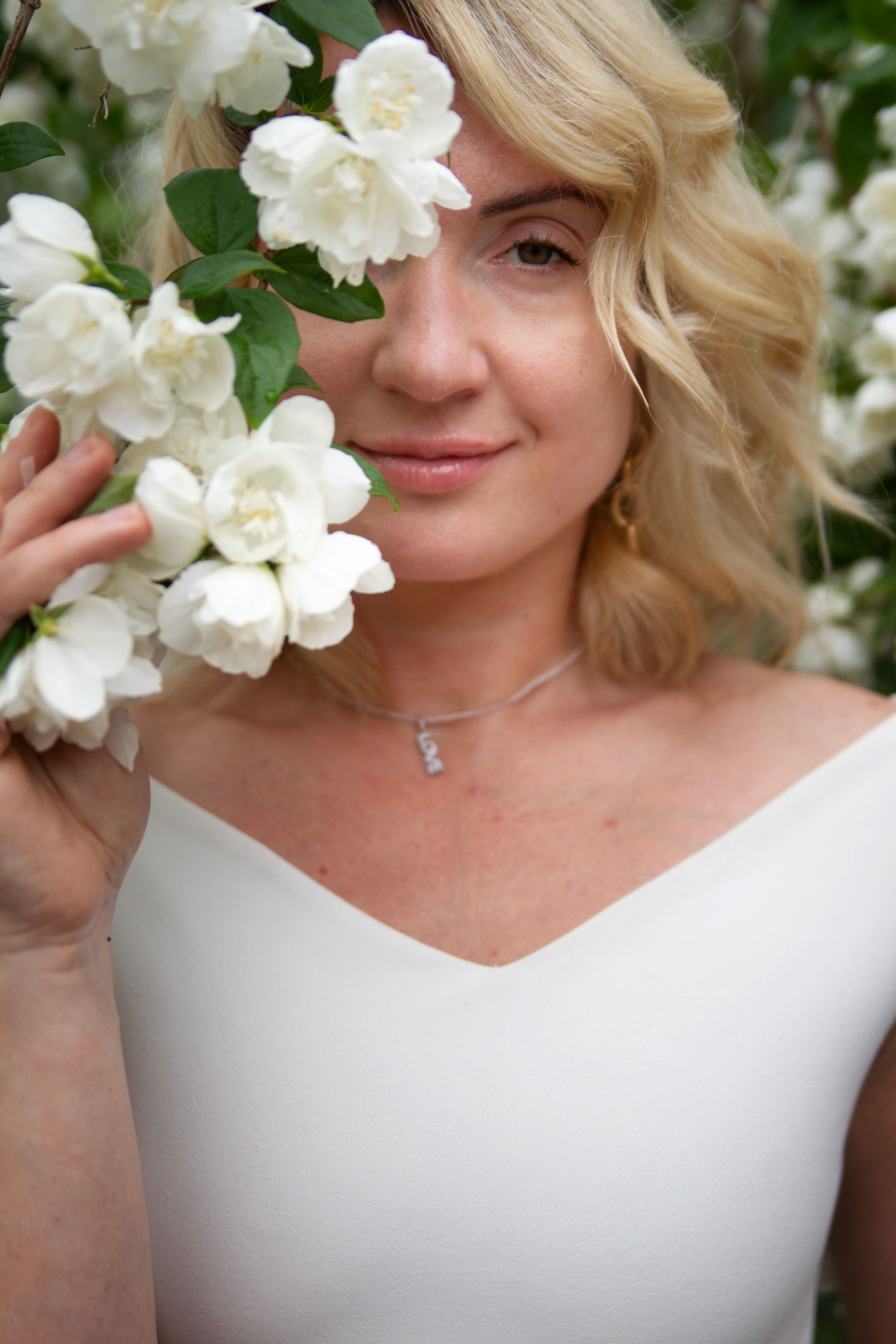Elegant blonde woman in white dress surrounded by blooming flowers, smiling gently.