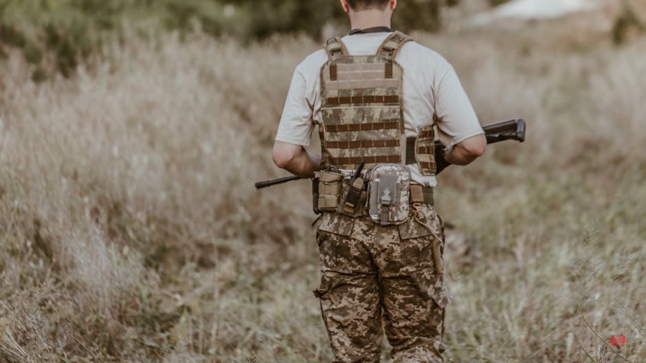 A back view of a soldier in camouflage uniform carrying a rifle in a grassy outdoor area.