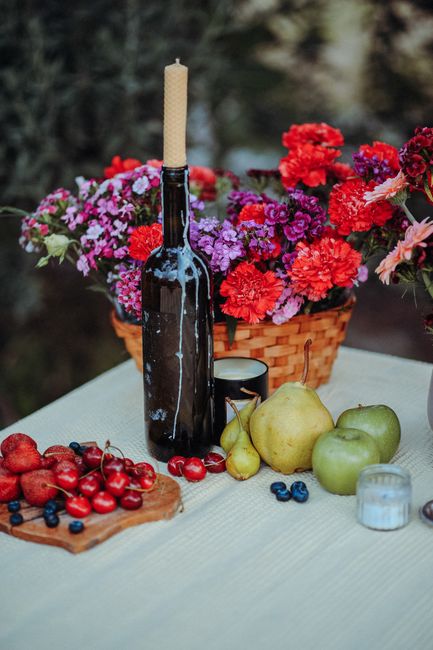 A colorful still life depicting fruits, a candle, and flowers on a table outdoors.