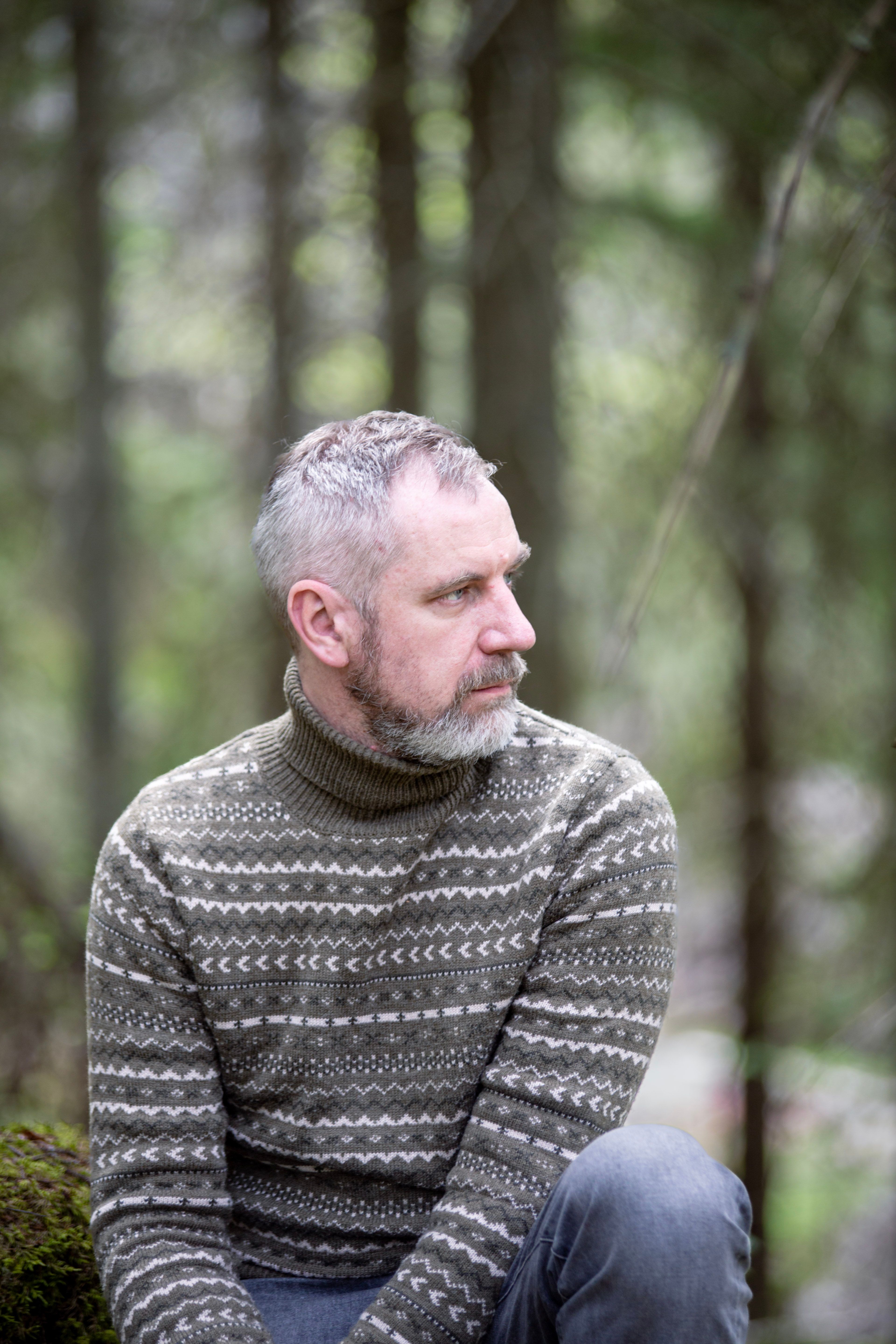 Pensive middle-aged man with beard and gray hair, wearing a patterned sweater, sitting in a forest.
