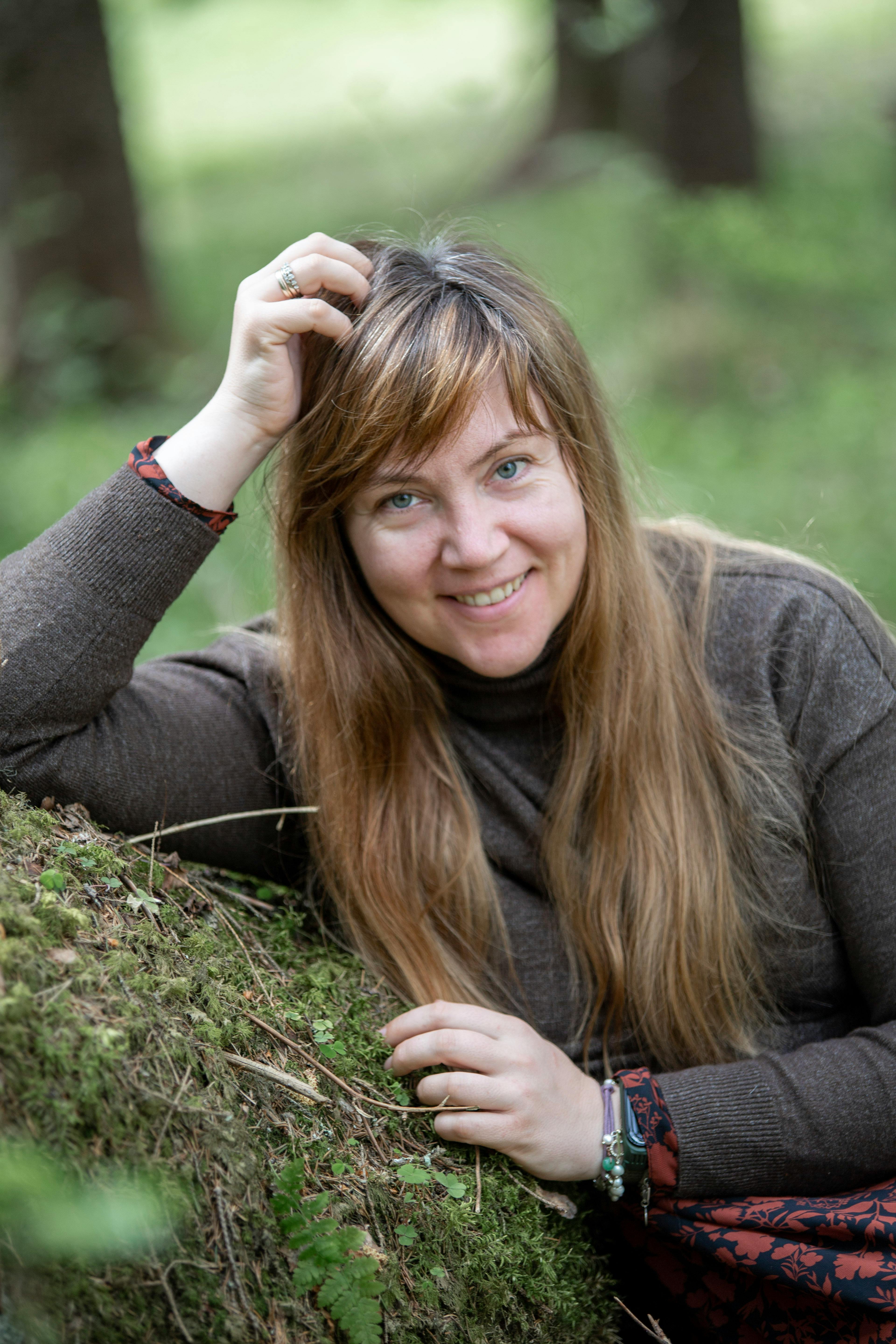 Portrait of a woman with long brown hair, smiling in a lush forest.