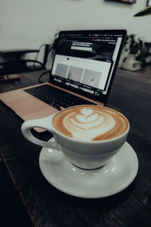 Free A coffee cup with latte art on a desk beside an open laptop in an office setting. Stock Photo
