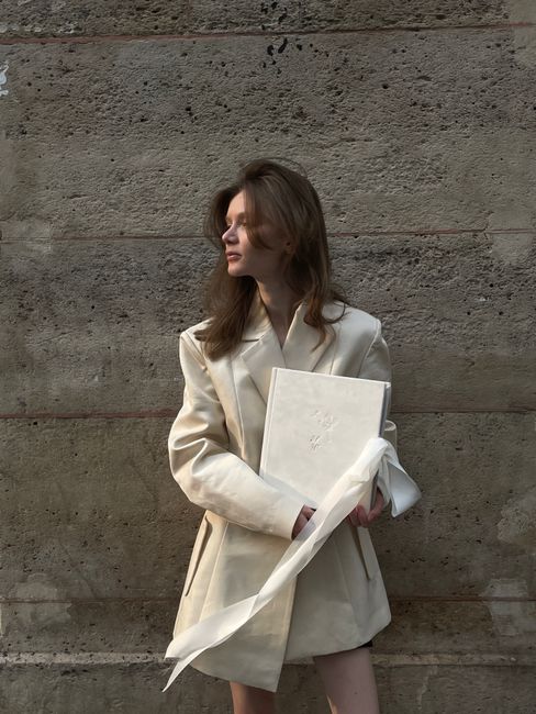Stylish woman in a white coat holding a book, standing against a textured wall.