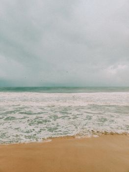 A tranquil view of an overcast beach with gentle waves meeting the sandy shore.