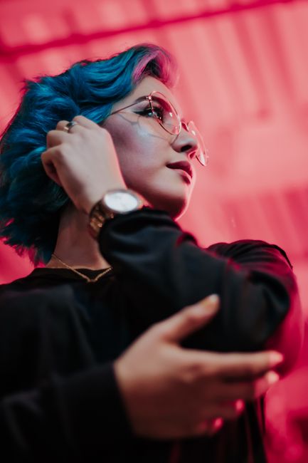 Portrait of a fashionable young woman with blue hair wearing glasses against a vibrant red background.
