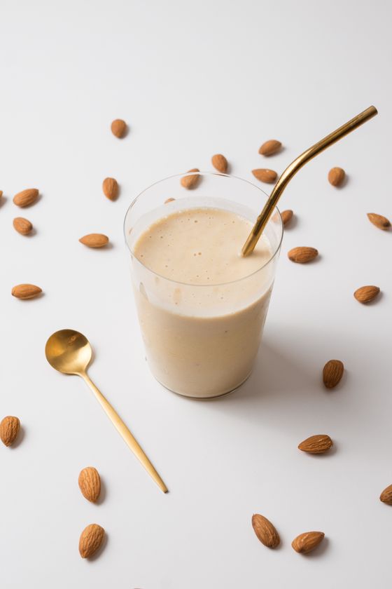 Beige almond smoothie in a glass with a gold straw and spoon among scattered almonds