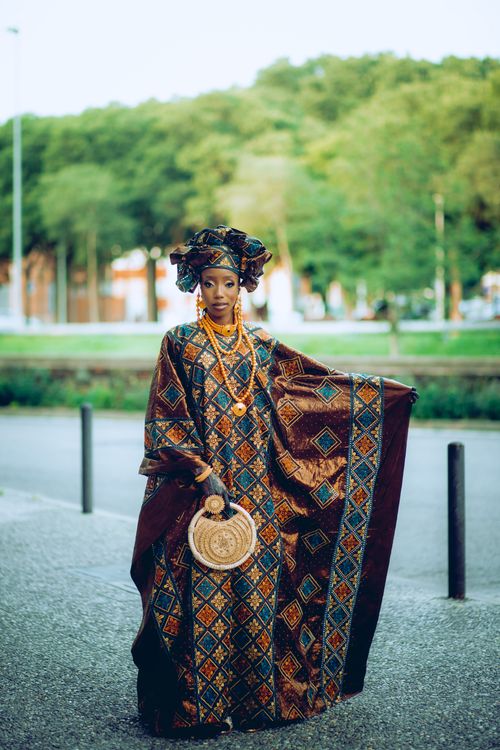 Free A young African woman poses gracefully in a vibrant traditional dress with a matching turban outdoors. Stock Photo