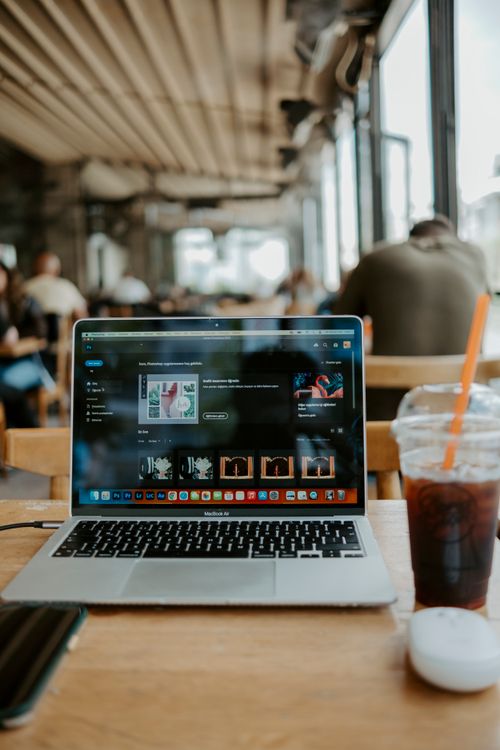 Free A laptop and iced coffee on a wooden table in a bustling cafe, ideal for remote work. Stock Photo