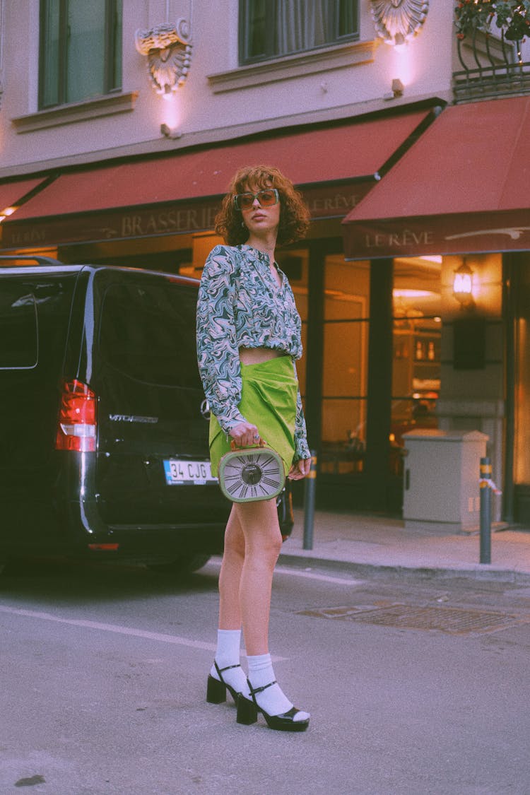 Young Woman In Abstract Pattern Blouse And Green Skirt Posing On A City Street At Dusk