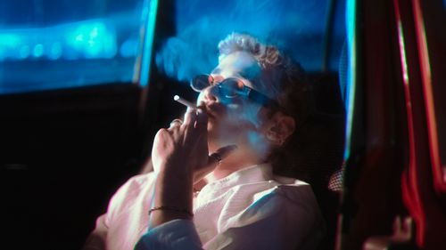 Free A man in sunglasses smokes a cigarette inside a car at night under blue neon lighting. Stock Photo