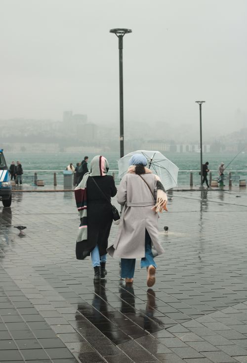 Free Two women walk with umbrellas on a rainy city day near a river. Stock Photo