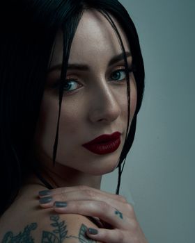 Close-up portrait of a woman with striking makeup and tattoos, captured in a dimly lit studio setting.