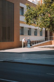 Two elderly men talking on a city street corner in Valencia under sunlight.