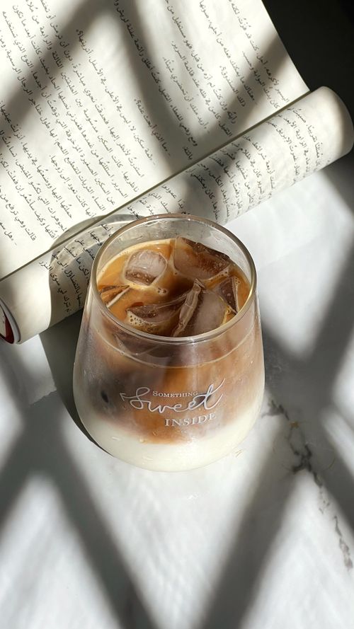 Free Glass of iced coffee on a sunlit table with Arabic script, offering a refreshing ambiance. Stock Photo