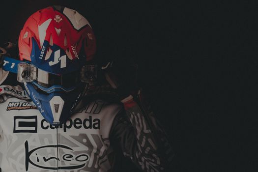 Professional motocross rider in Italy, wearing vibrant helmet and racing suit, posing confidently in dark setting.