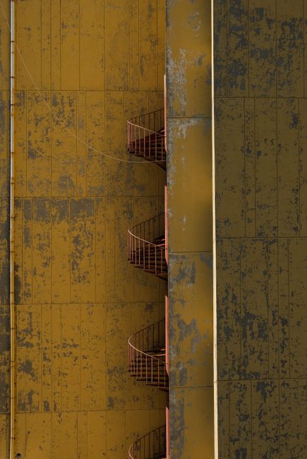 Vertical shot of a rusty spiral staircase on a yellow industrial building facade.