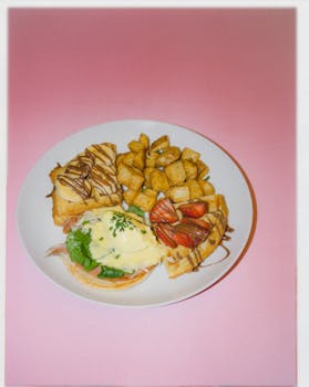 A beautifully arranged brunch platter featuring waffles, eggs benedict, and fries on a pastel pink background.