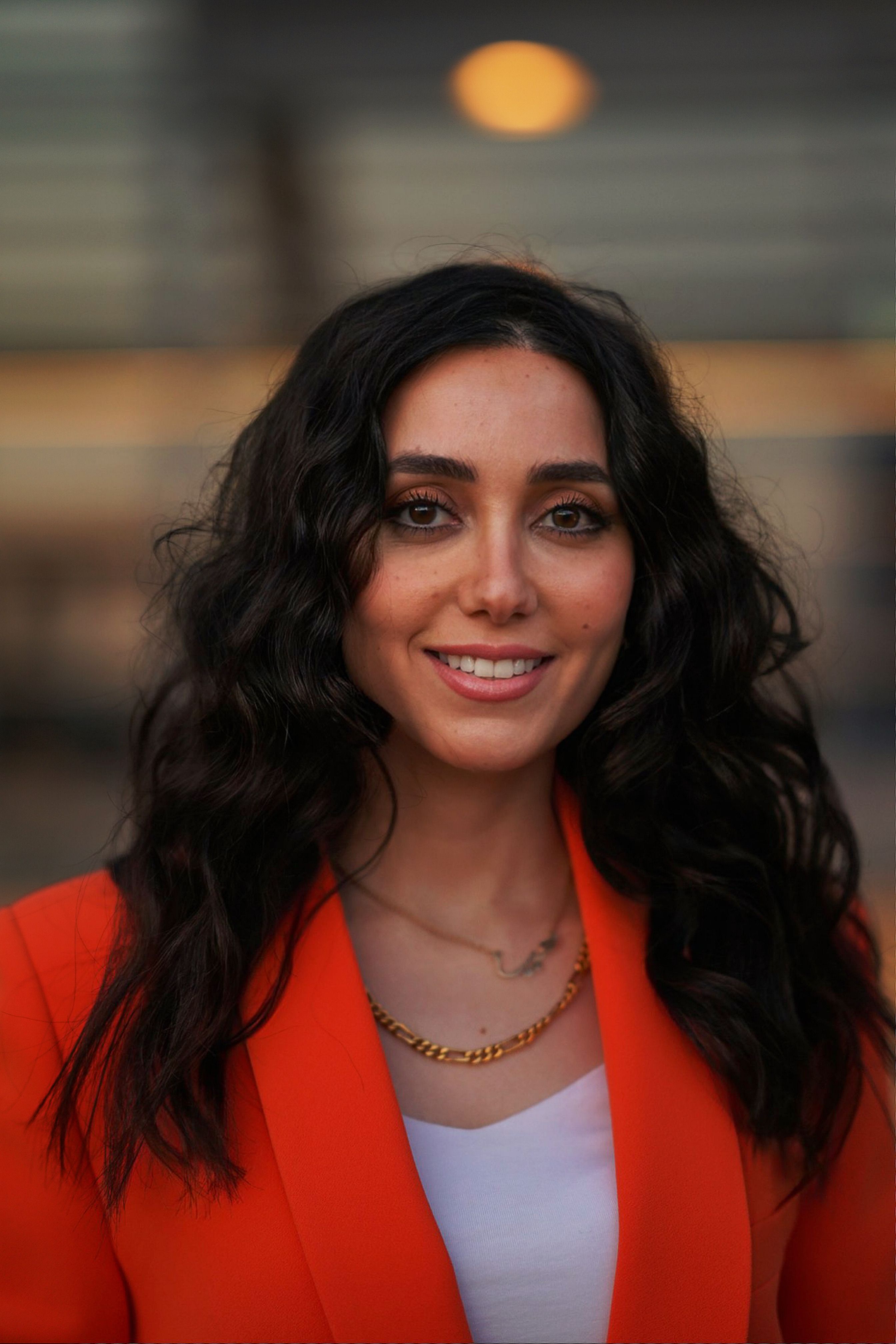 Elegant woman with dark hair and orange jacket smiling confidently outdoors.