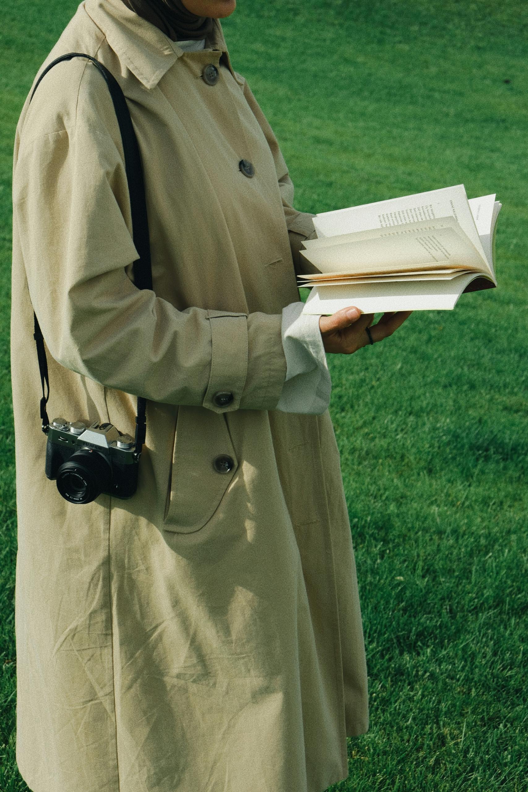 A woman reading a book in a trench coat with a camera in a park setting. Captured outdoors.