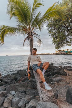 A young man sits by a palm tree on the rocky coast, enjoying the ocean view.