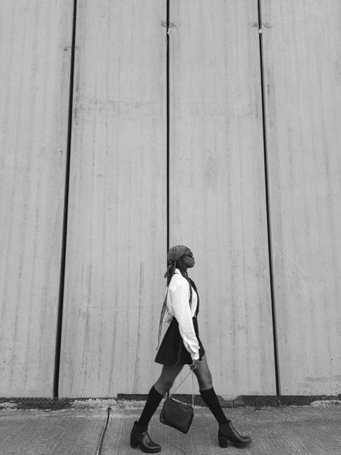 A woman walking stylishly by a wall, captured in a black and white urban setting.
