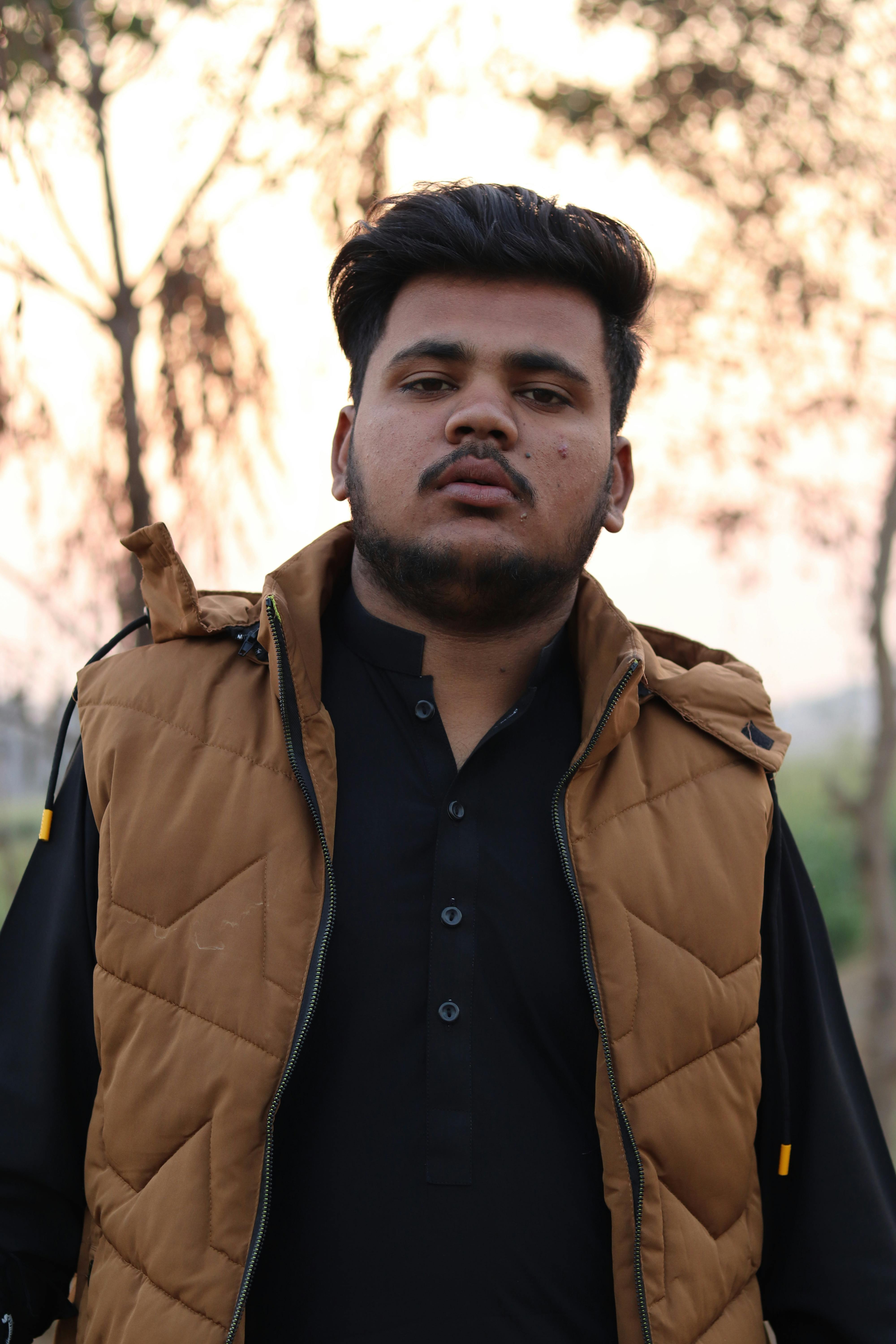 Portrait of a young man with black hair wearing a tan vest, standing outdoors during the day.