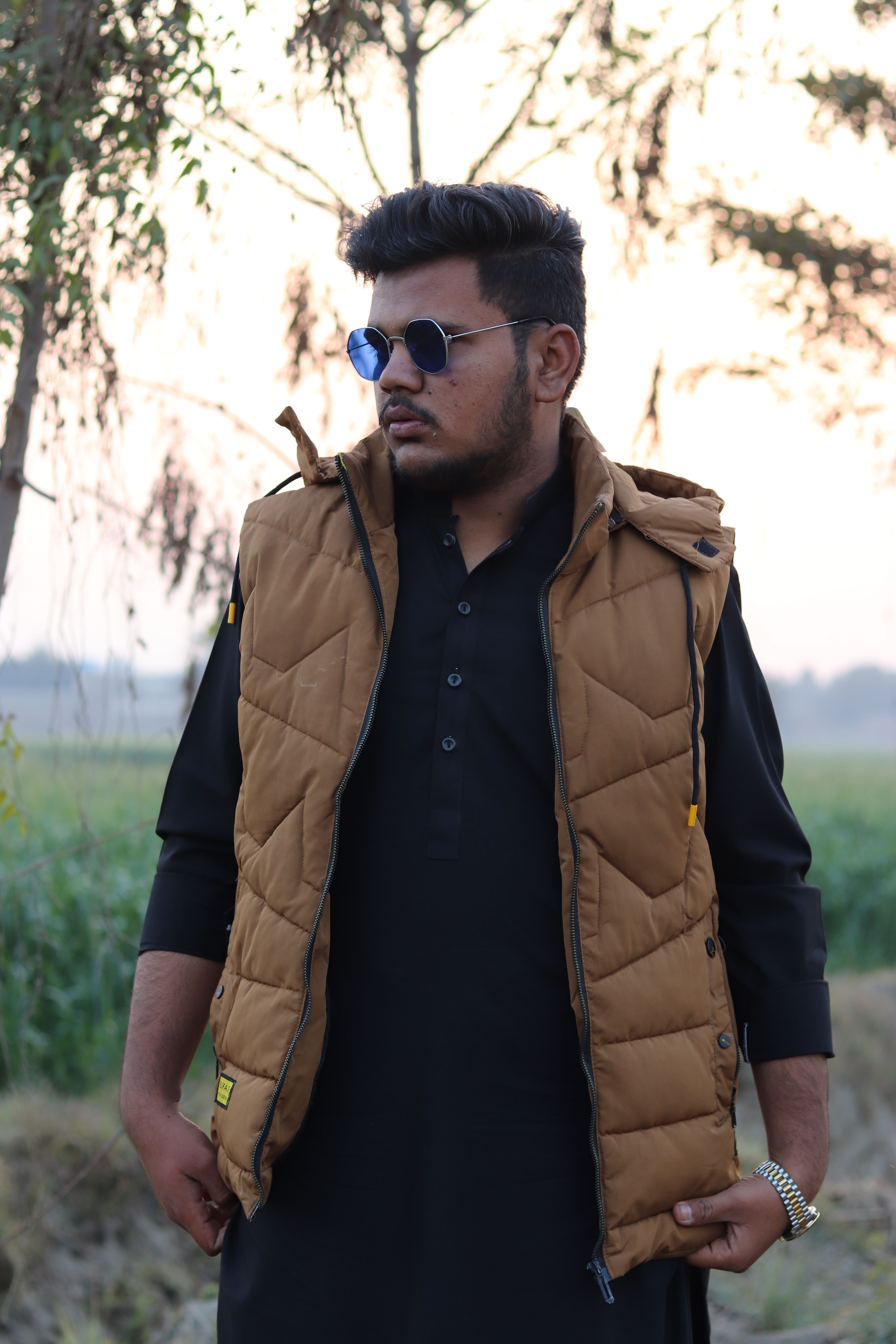 Young man wearing a brown vest and sunglasses standing outdoors, exuding confidence.