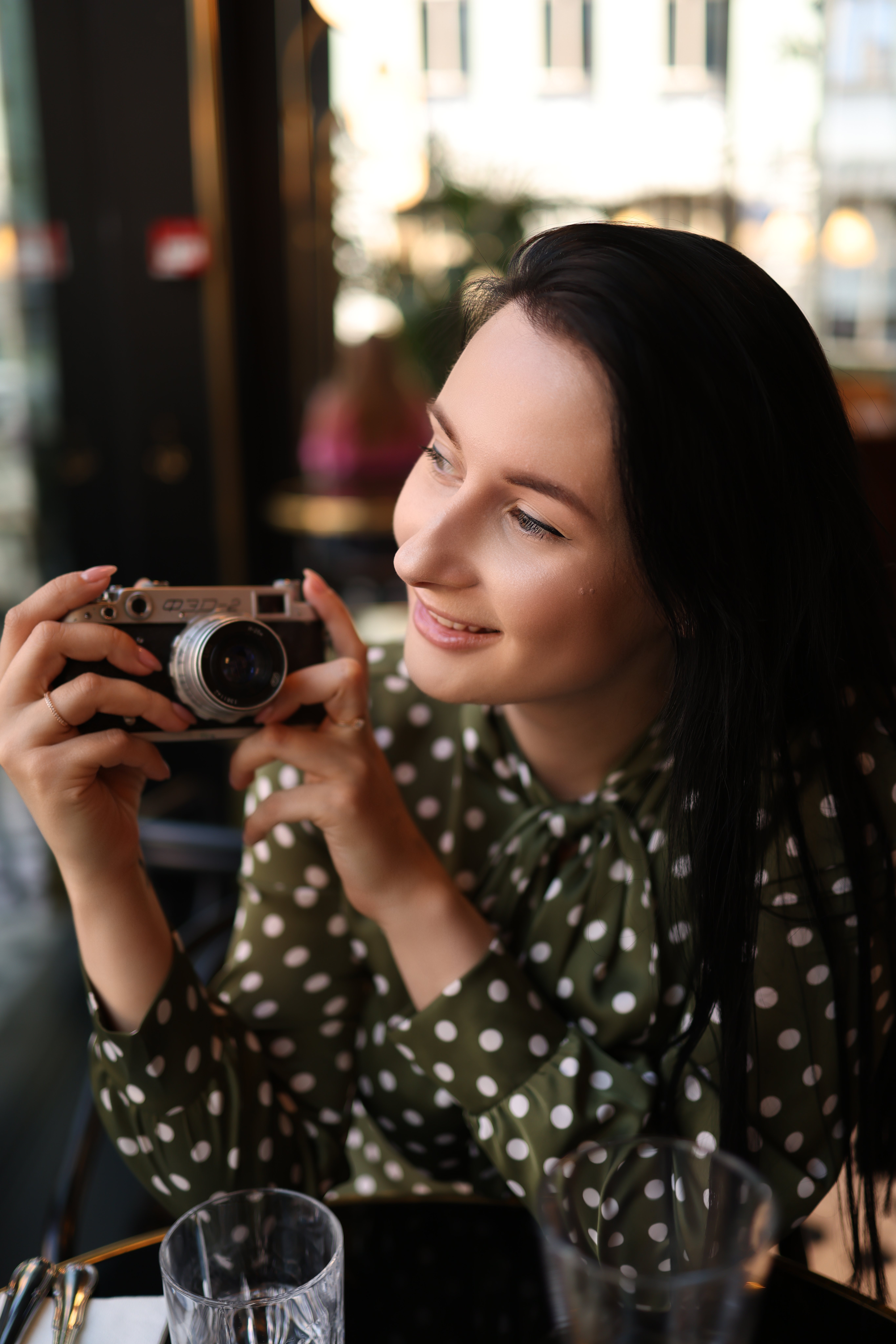 Smiling woman in polka dot dress holding a vintage camera indoors.