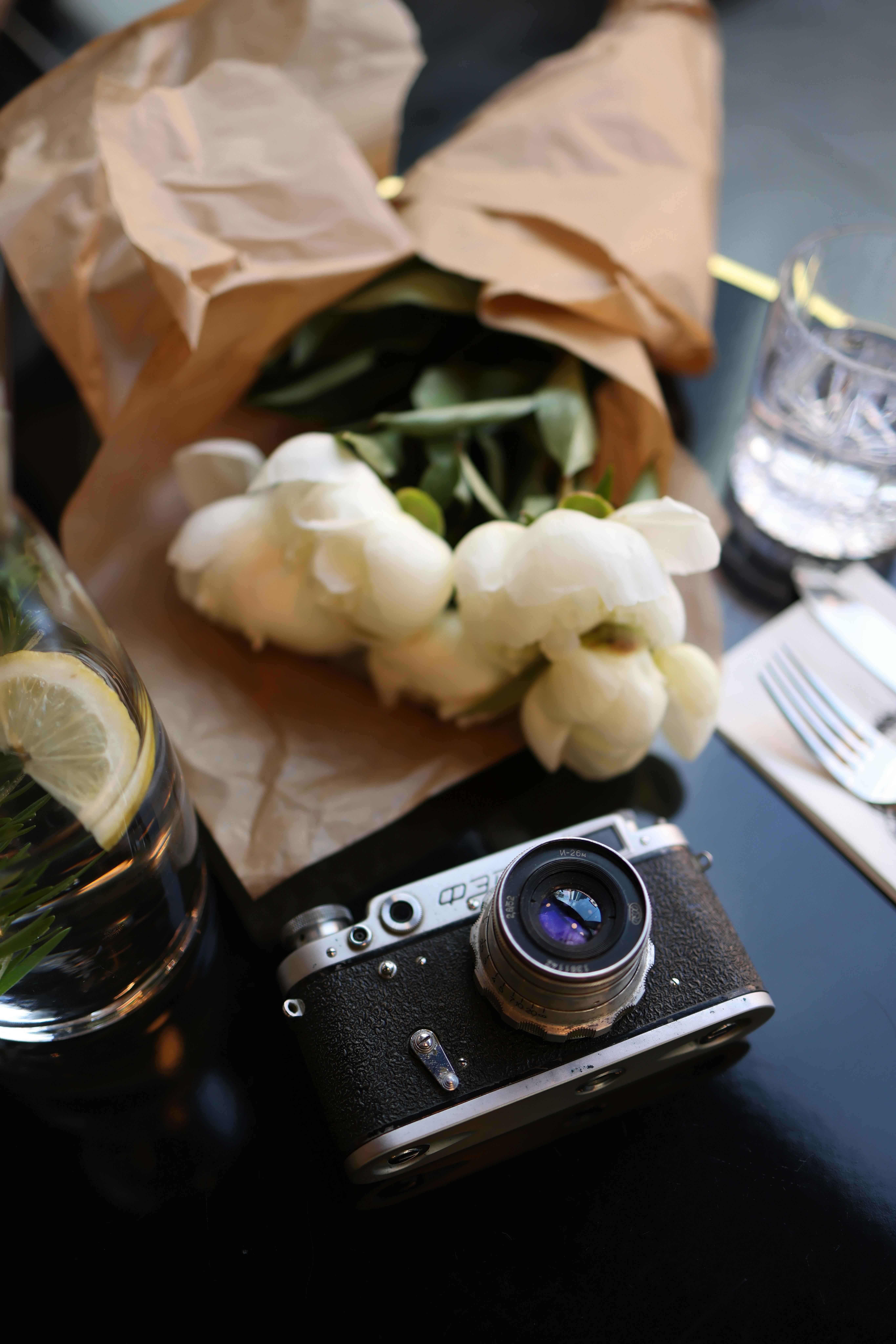 A vintage camera alongside white tulips wrapped in brown paper, captured on a reflective surface.