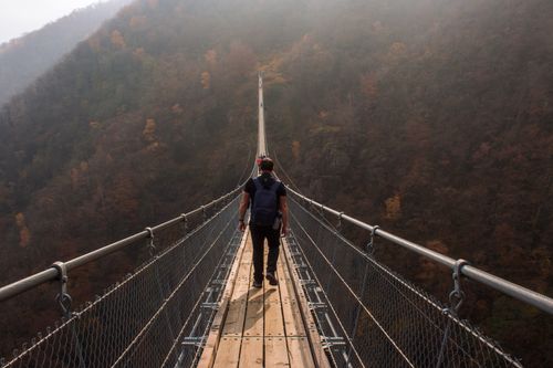 Free A person hikes across a long suspension bridge surrounded by fall foliage. Stock Photo