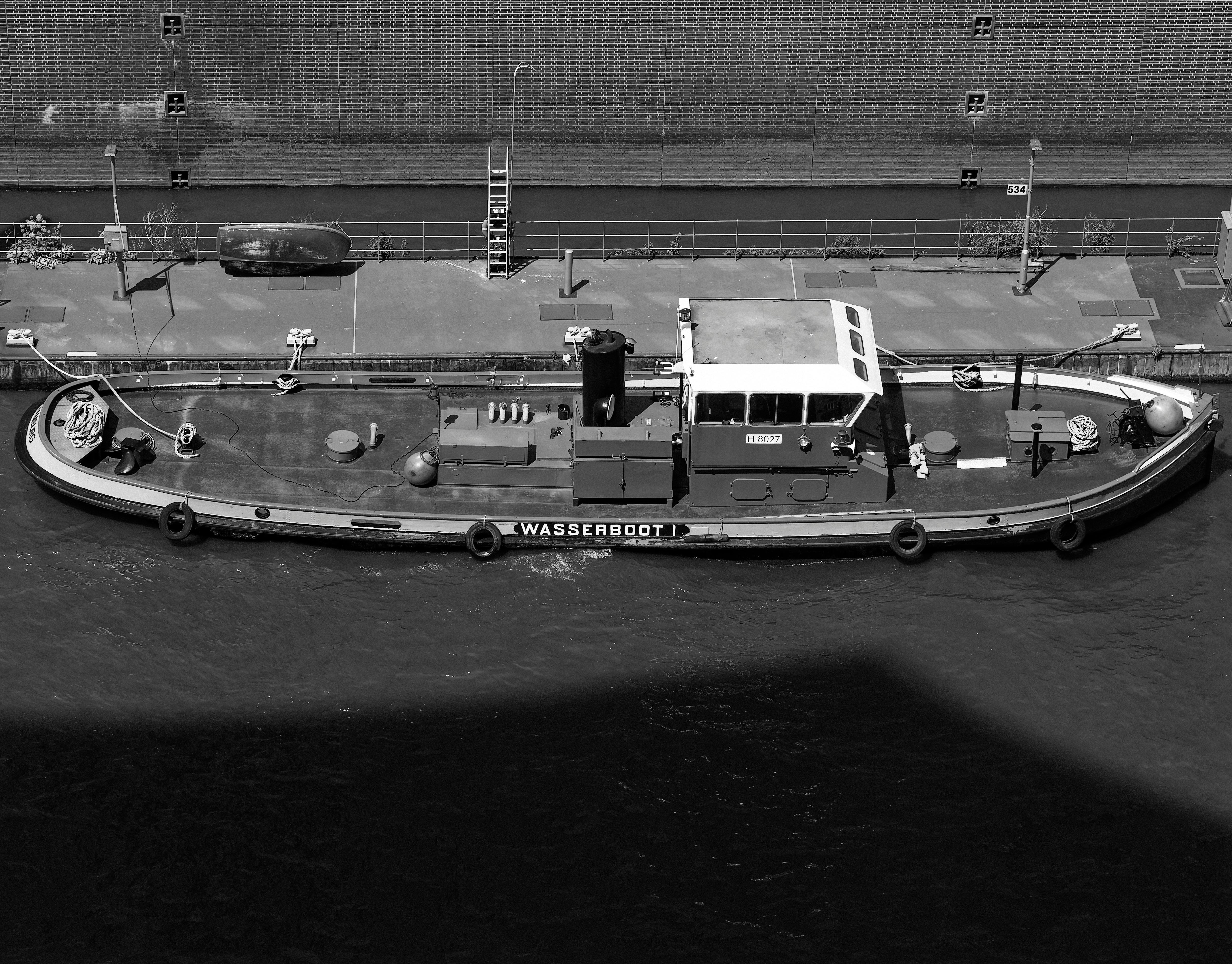 Black and white aerial photo of a moored tugboat on a city waterway with a concrete wall in the background.