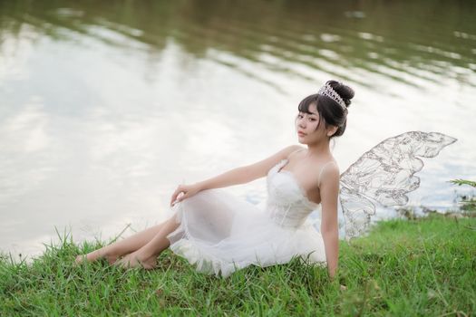 Asian woman in fairy cosplay sitting by the water in a mystical setting, evoking a dreamlike atmosphere.