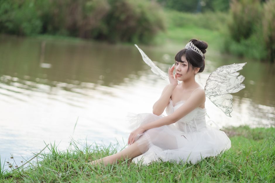 A woman dressed as a fairy sits gracefully by a riverside, capturing a sense of whimsical elegance.