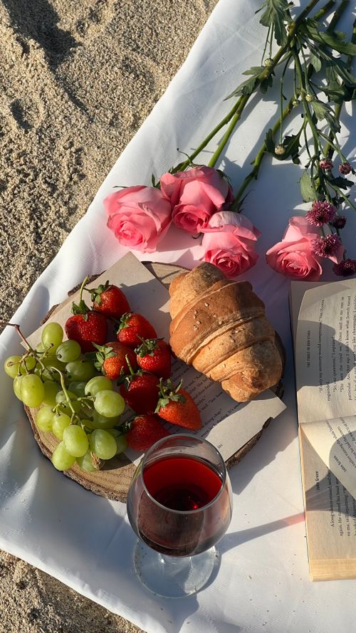 Free A beautiful beach picnic scene featuring roses, a croissant, fruits, and a glass of red wine. Stock Photo