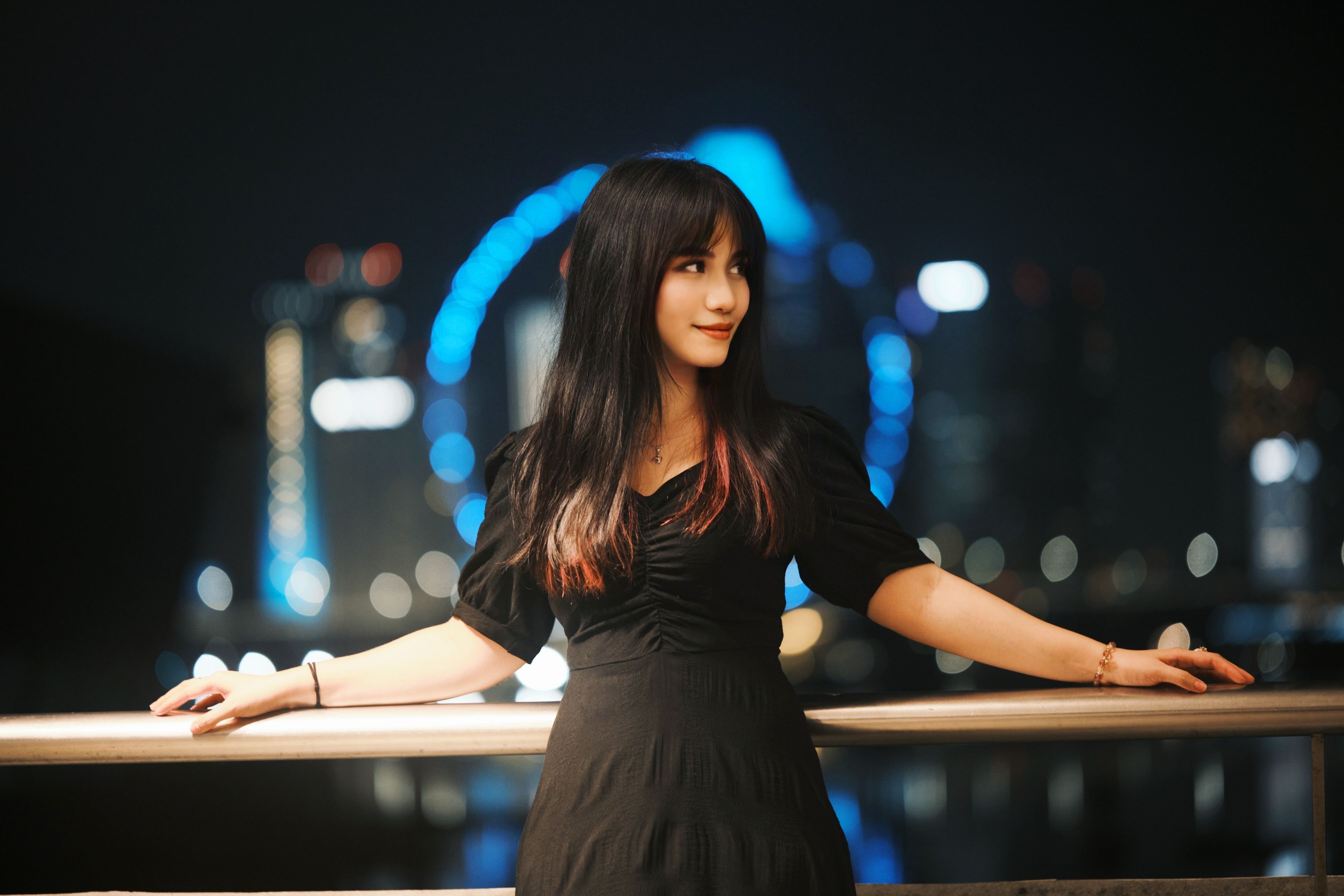 Fashionable woman in black dress posing by handrail with city lights in the background.