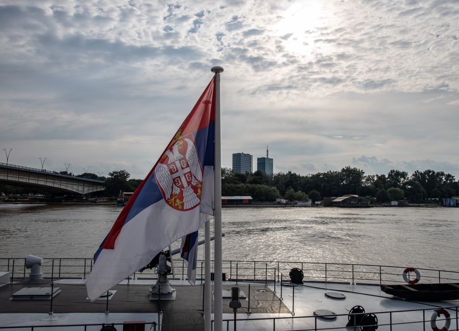 Serbian flag on a boat with an urban skyline and river scene in daytime.