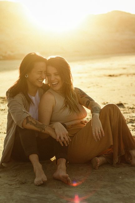Two women laughing and hugging on a sandy beach during sunset.