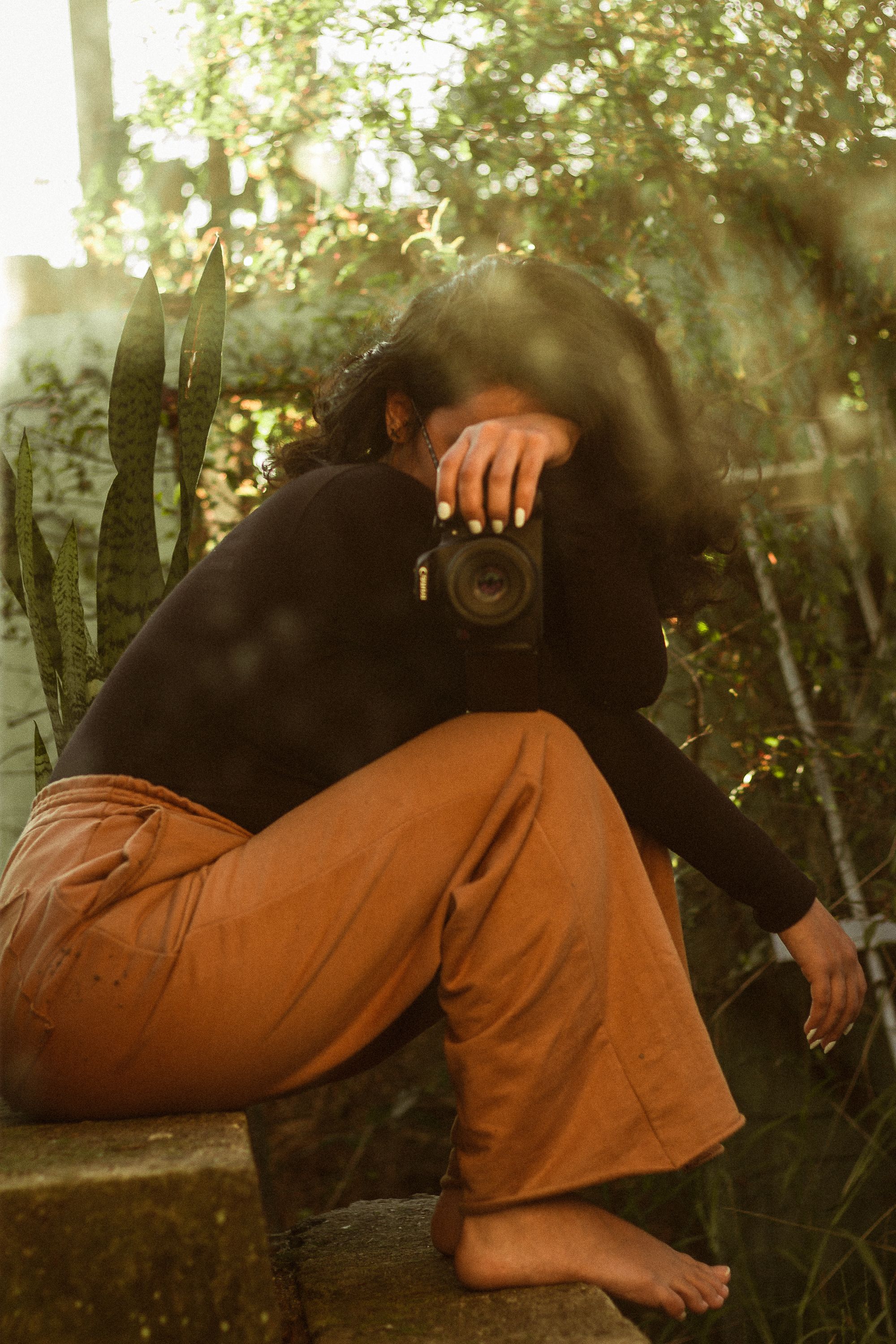 A woman with brunette hair sits barefoot outdoors, holding a camera on stairs surrounded by greenery.