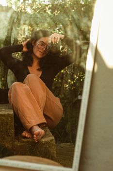 Woman sitting on stairs uses a camera to capture a mirror reflection in warm, outdoor setting.