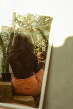 A woman is seated on stairs using a camera to capture a self-portrait in a mirror reflection outdoors.