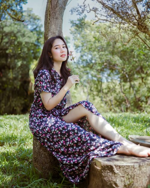 Free Young woman sitting on a tree stump in a floral dress during a sunny day in nature. Stock Photo
