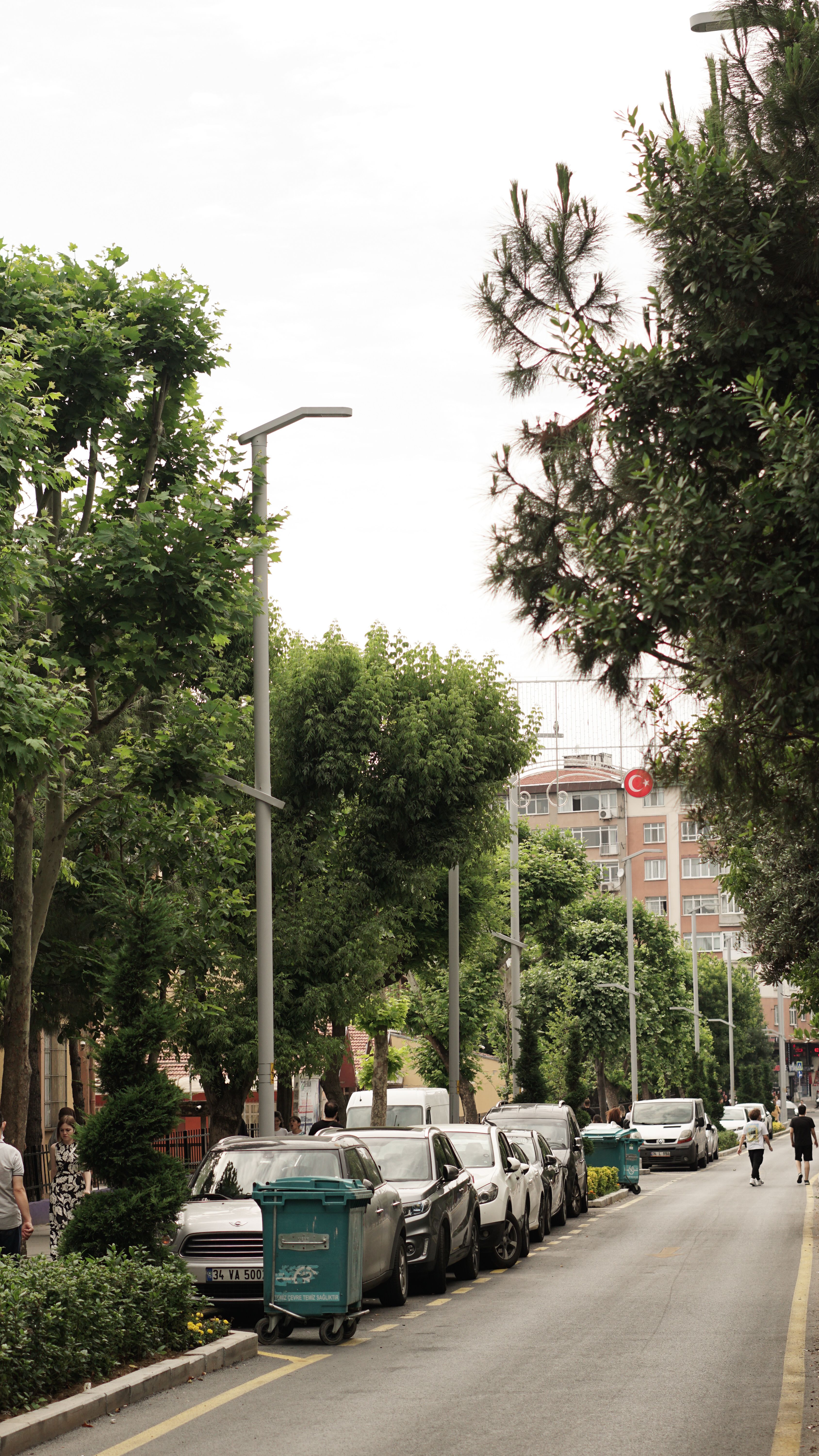 A quiet city street lined with trees, cars parked along the sides, and pedestrians walking.