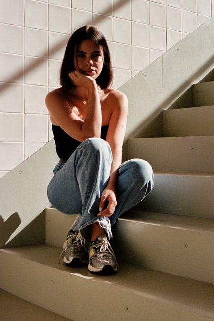 A woman in casual attire sits thoughtfully on stairs, lit naturally indoors.
